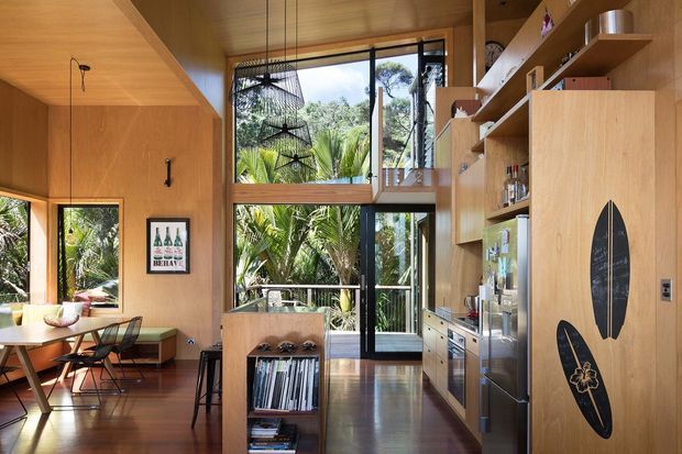 Warm timber-lined open-plan interior with a dining area on the left and a compact kitchen and built-in shelving on the right. A tall glazed opening and glass door bring in abundant natural light and overlook lush tropical vegetation. The space features polished timber flooring, extensive timber cabinetry and wall finishes, open shelving, a built-in dining bench, and surfboard-themed decorative panels.