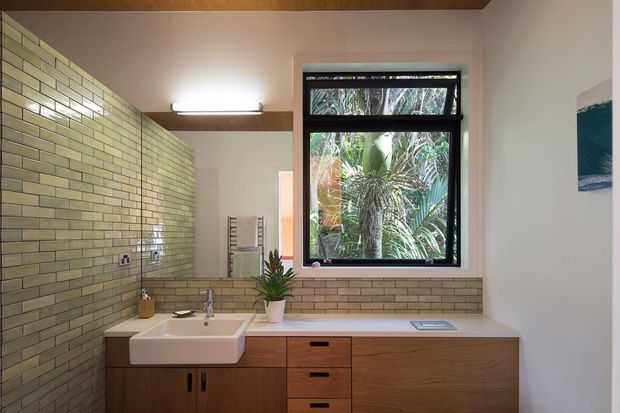 A warm, contemporary bathroom vanity area with a long timber cabinet, white countertop and inset basin, wall-mounted chrome tap, and extensive pale green glazed brick wall tiling. A black-framed window overlooks dense tropical planting, while a timber-lined ceiling and simple linear wall light add warmth. A small artwork is visible on the right wall.