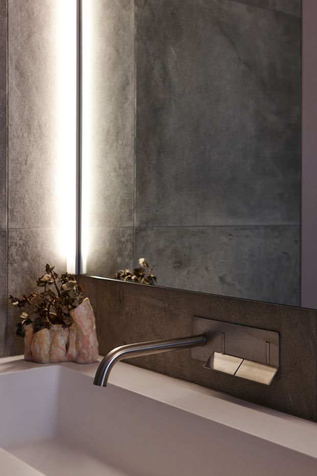Close-up view of a contemporary bathroom vanity area with a deep rectangular basin, wall-mounted brushed metal tapware, and a recessed wall-mounted control or soap fixture. Large dark grey textured stone-look wall tiles form the backsplash, with a narrow vertical illuminated mirror or light feature beside the basin. A sculptural decorative vase with dried foliage sits on the ledge, adding a warm natural accent.