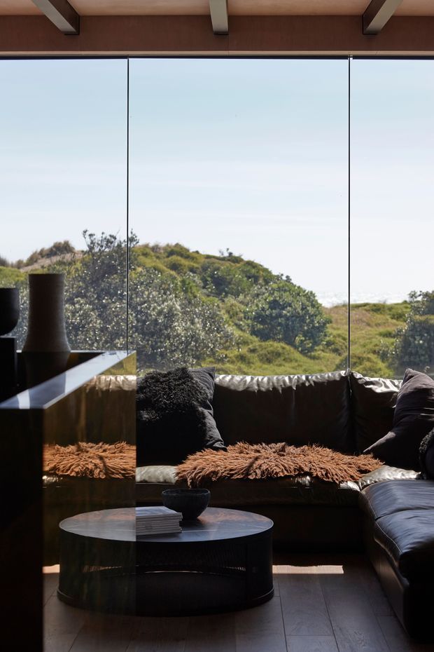 A sophisticated living room with a dark leather sectional sofa, textured brown throws and cushions, a low round coffee table, and sculptural dark decor. Full-height glazing provides expansive views toward dense coastal vegetation and bright sky. The room has a warm timber ceiling with exposed structural beams, dark reflective built-in joinery, and dark timber flooring.