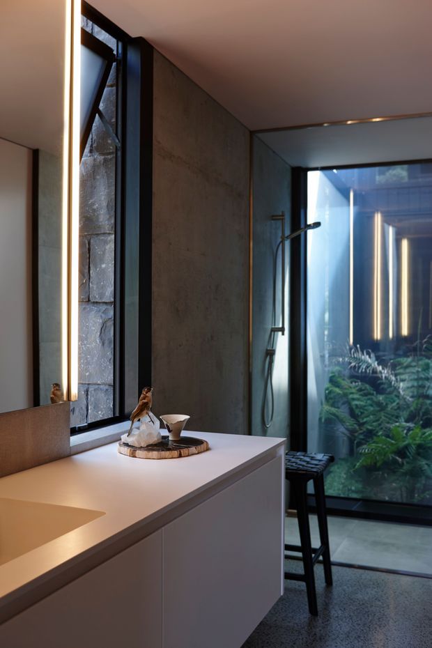 A refined contemporary bathroom interior featuring a long white floating vanity with integrated basin, large illuminated mirror, dark-framed window, concrete-look wall surfaces, and a frameless glass shower enclosure. Warm linear lighting, black tapware and hardware, terrazzo-like flooring, and indoor greenery visible beyond the glazing create a calm, luxurious atmosphere.