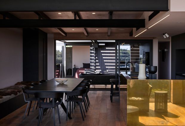 Dark, sophisticated open-plan interior combining a dining area, living room and kitchen. A large black dining table with multiple chairs occupies the foreground, while a black sofa and coffee table sit beyond it near full-height glazing. The kitchen features a reflective golden island or benchtop, dark cabinetry, a sink and tap, and a white sculptural vase. Exposed dark structural beams, timber ceiling panels, recessed lighting and linear LED lighting contribute to a dramatic contemporary aesthetic.