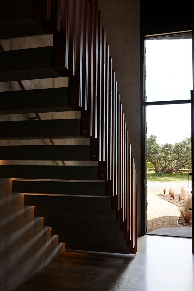 A dramatic interior view of a contemporary stairway with dark open-tread stairs and a vertical timber balustrade forming a stepped rhythm alongside a textured wall. A tall black-framed glazed door or window opens toward a landscaped exterior, while daylight creates strong contrast against the shadowed stairwell. The floor appears smooth and polished, and the overall composition is minimalist and architectural.