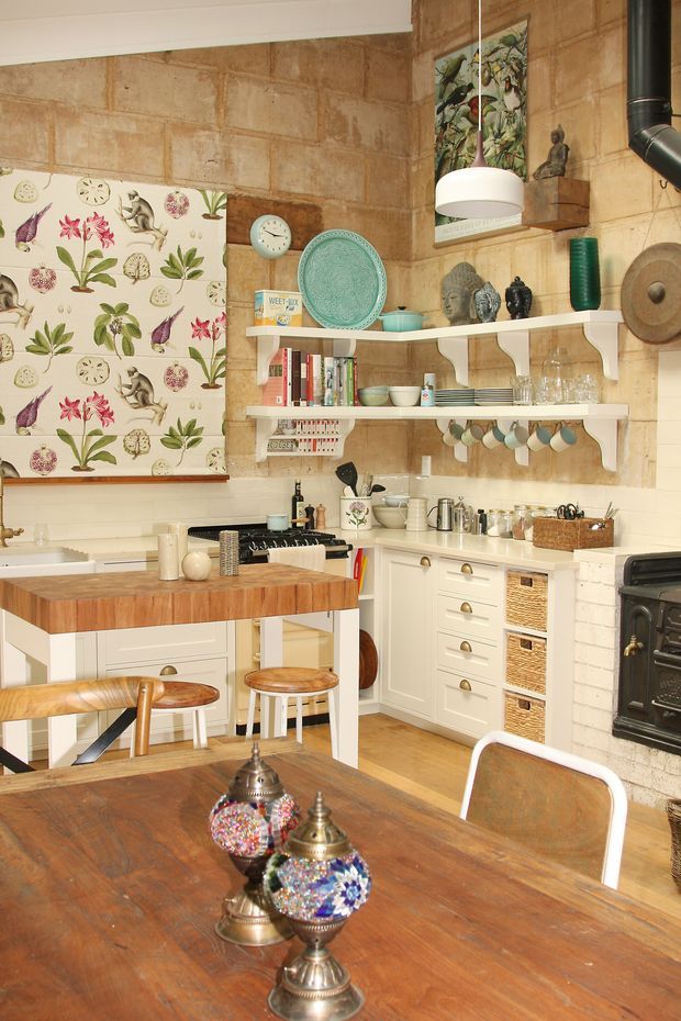 A warm, eclectic kitchen and dining area with exposed tan brick walls, white cabinetry, open shelving, patterned botanical window covering, and a timber-topped island with stools. A dark vintage-style range cooker, hanging pendant light, crockery, books, decorative objects, and a large rustic timber dining table create a homely, collected look.