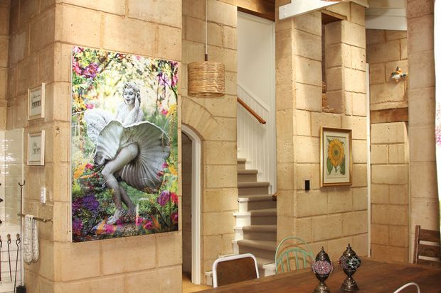 Interior view of a warm, eclectic dining and entry area with pale sandstone block walls, a timber dining table, decorative artwork, and a staircase rising through the centre of the space. A colourful floral artwork and framed botanical print add vivid accents, while woven and glass decorative elements contribute to the layered styling.