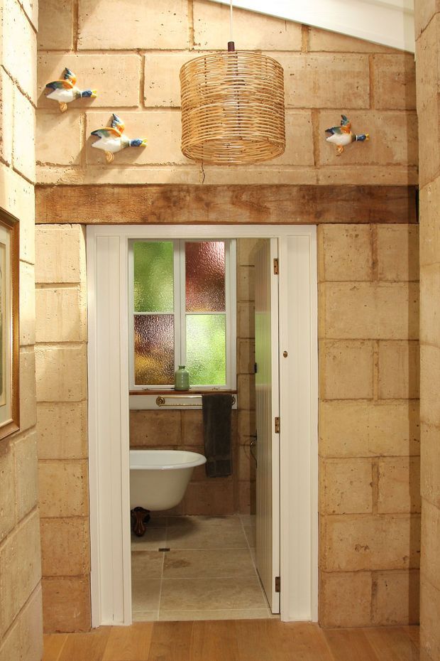 A warm, rustic interior hallway or entry area with exposed tan stone block walls, a heavy timber lintel, and timber flooring. A white-framed doorway opens directly into a compact bathroom containing a freestanding-style white bathtub, tiled floor, textured coloured glass window, and dark towel. A woven rattan pendant light and decorative bird wall mounts add character.