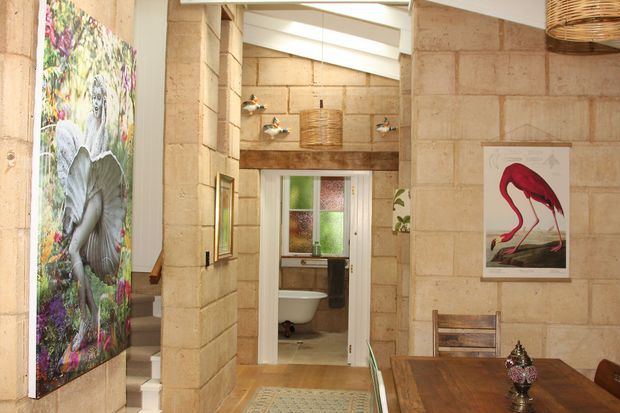 Warm, eclectic interior view through a hallway toward a bathroom. The space features exposed light stone block walls, timber flooring and joinery, white structural beams, woven pendant lighting, colourful botanical and flamingo artwork, and a timber dining table in the foreground. A freestanding bathtub and patterned glazing are visible in the bathroom beyond.