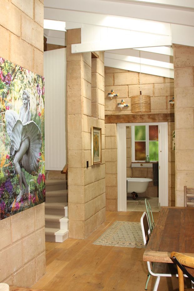 Interior view of an open-plan dining and circulation area with exposed tan masonry block walls, timber flooring, a timber dining table and chairs, and a stairway. A bathroom is visible through a doorway, along with colourful artwork, decorative wall lights, and a woven pendant light. White structural beams and high-level glazing create a bright, airy space.