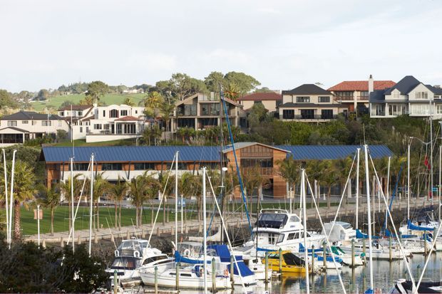 Wide exterior view across a marina with numerous sailboats in the foreground, a low waterfront building with a blue metal roof and timber-toned facade in the middle ground, and detached multi-storey houses on a gently sloping residential hillside behind it. The scene is photographed outdoors in daylight with a coastal waterfront setting.