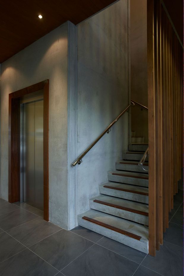 A dimly lit interior stairway with exposed concrete walls and concrete stairs accented by dark timber nosings. A slim metal handrail follows the stair, while vertical timber slats form a screen along the right side. A stainless-steel elevator with a dark timber surround is visible on the left. The space has a dark timber ceiling, recessed lighting, and large dark grey tiled flooring, creating a restrained, materially focused architectural interior.