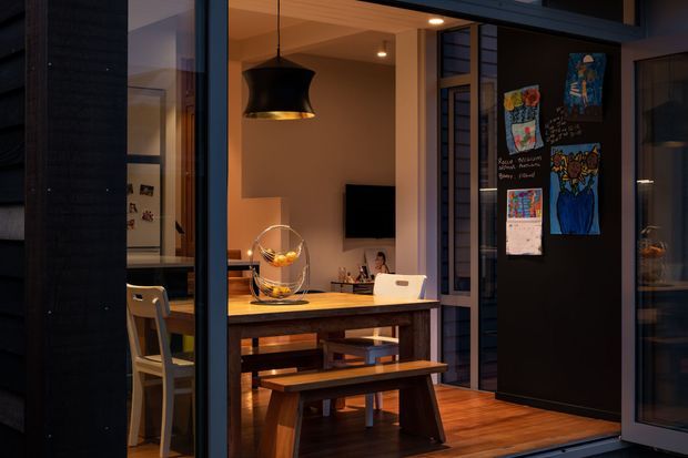 Warmly lit contemporary dining and kitchen-adjacent interior viewed through dark-framed glazing. A timber dining table with bench seating, timber flooring, pendant lighting, exposed ceiling beams, a wall-mounted television, and a dark feature wall displaying children's artwork are visible. The space has strong indoor-outdoor glazing and a compact, family-oriented atmosphere.