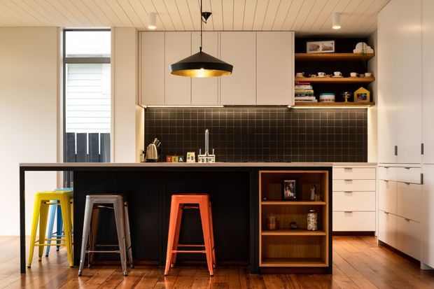 Warm contemporary kitchen with a central island featuring a dark waterfall-style work surface, open shelving, and three colourful stools. The kitchen includes pale flat-panel cabinetry, a black square-tile splashback, timber flooring, integrated white storage, and a sculptural black pendant light. A narrow glazed door or window provides additional daylight, while open timber shelving displays crockery and decorative objects.