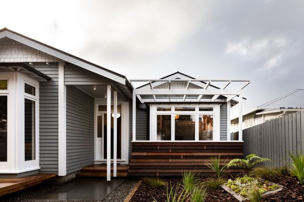 Exterior view of a contemporary coastal-style home featuring grey horizontal weatherboard cladding, white-trimmed windows, a pitched gabled roof, covered entry, timber decking, and a landscaped front garden. A low timber privacy screen and white pergola-like structure define the outdoor area.