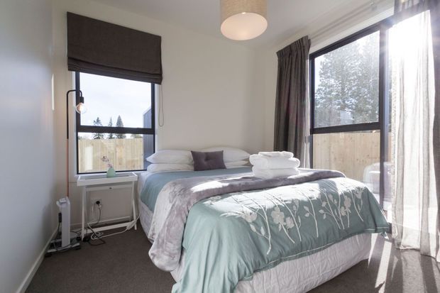 Bright, compact bedroom with a neatly made bed dressed in pale blue-green floral bedding, layered white pillows, folded towels, and a soft grey throw. The room has white walls, dark carpet, roller blinds and full-height curtains, large windows, a simple bedside table, and a pendant light. Natural daylight creates an airy, calm atmosphere.