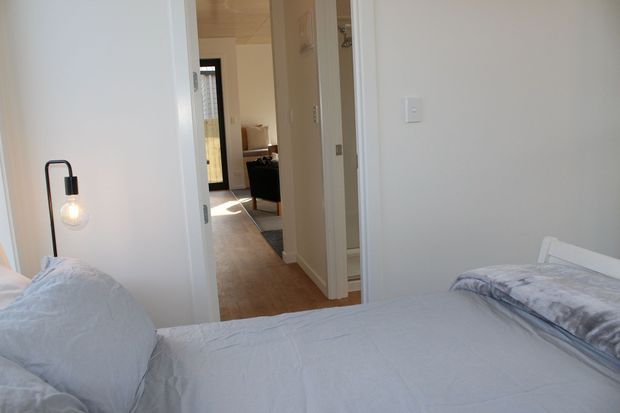 A bright, simply furnished bedroom with two single beds or a twin-bed arrangement in the foreground, pale bedding, white walls, and a black minimalist bedside lamp. An open doorway leads to a hallway and a partially visible living area beyond. The space has a clean, contemporary feel with timber flooring and abundant natural light.