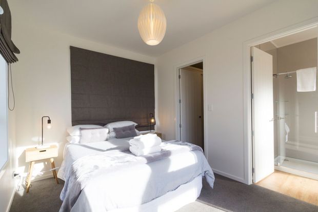 Bright, compact contemporary bedroom with a neatly made upholstered bed, layered white and pale grey bedding, and a large dark textured rectangular feature headboard. A warm pendant light, slim bedside lamps, and a small timber bedside table add understated detail. White walls and trim, carpeted flooring, and an open doorway to a tiled bathroom are visible.