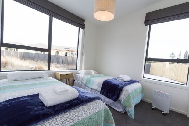 Bright twin bedroom with two single beds dressed in coordinated aqua, white, and navy bedding, folded towels, dark carpet, large black-framed corner windows, roller blinds, a small timber bedside table, and a simple pendant light. The room has a clean, practical, contemporary feel.