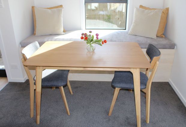 Bright, compact dining nook with a light timber rectangular dining table and four upholstered wooden chairs. A built-in window seat with neutral cushions sits beneath a large window, while dark grey carpet and white walls create a clean, calm interior.