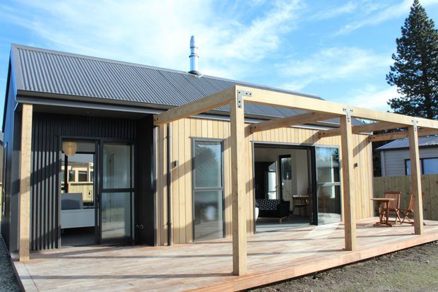 Single-storey contemporary residential building viewed from the exterior, with dark corrugated metal cladding and roofing, light timber-clad walls, large glazed doors and windows, and a broad timber deck. An open timber pergola frames the deck and entry, while a visible chimney suggests an internal wood-burning stove. The image shows a sunny, open outdoor living area and portions of the interior through the glazing.