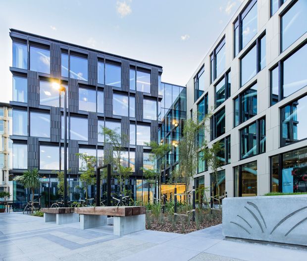 Contemporary multi-storey commercial or mixed-use building complex arranged around a landscaped paved courtyard. The facades combine expansive blue-tinted glazing, dark vertical cladding panels, white framing and concrete elements, with benches, planters, young trees and outdoor seating in the foreground.