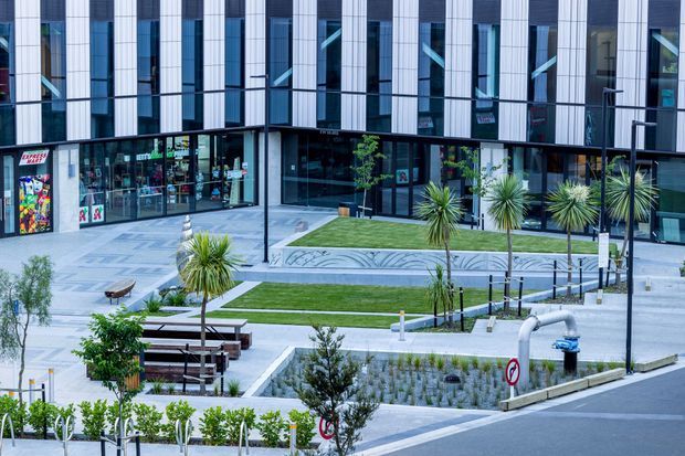 Contemporary multi-storey commercial building with a highly glazed ground-floor frontage, vertically articulated white facade panels, and dark window bands. The landscaped forecourt includes paved walkways, raised lawn areas, young trees, palms, bench seating, and integrated stormwater planting beds.