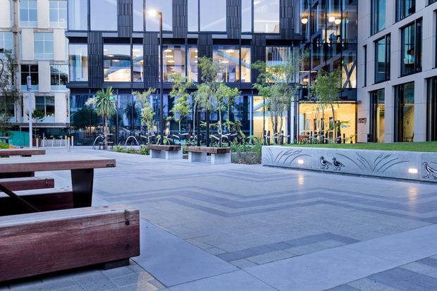 A landscaped urban courtyard or public plaza framed by contemporary multi-storey buildings with extensive glazing, dark vertical facade elements, planted raised beds, timber benches, paved walkways, and an integrated curved concrete seating wall with botanical-style detailing. The scene is photographed at dusk with warm interior lighting visible through the windows.