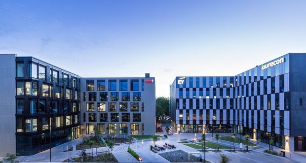 Wide exterior view of a contemporary multi-building commercial office precinct at dusk, with dark and silver-blue glazed facades, regular grid windows, landscaped pedestrian courtyards, paved paths and outdoor seating. The Aurecon-branded office building is prominent on the right, with additional modern office blocks surrounding the central plaza.