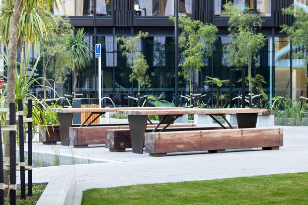 A contemporary landscaped outdoor courtyard or plaza in front of a dark glazed building, featuring a large timber communal table with integrated bench seating, planters, young trees, tropical planting, concrete paving, and lawn. The building has black vertical cladding, extensive glazing, and a modern commercial character.