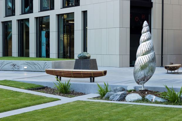 Landscaped courtyard or plaza outside a contemporary multi-storey building, featuring pale tiled facade panels, extensive glazing, manicured lawns, concrete paths and seating. Sculptural metal artworks and a shallow rock-lined water feature form focal points in the foreground.