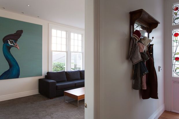 View from an entryway into a bright living room, featuring a dark sofa, low rectangular coffee table, carpeted floor, large white-framed windows, and a large bird artwork. The entry area has a traditional wooden coat rack with hanging garments and a decorative stained-glass sidelight beside a pink-toned door.