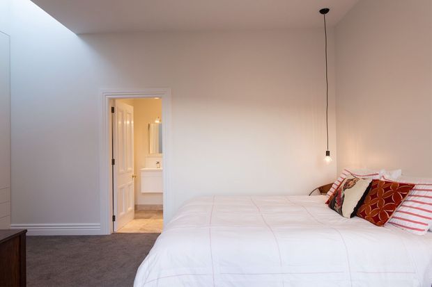 Bright, simply furnished bedroom with a large white bed, patterned and striped accent cushions, soft grey carpet, white walls and trim, and a minimal hanging pendant light. An open doorway leads directly to a bathroom with a visible vanity.