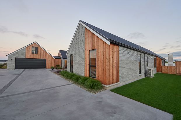 Exterior view of a contemporary single-storey residential complex with two gabled volumes. The buildings combine pale grey masonry walls with vertical natural timber cladding, dark metal roofing, narrow vertical windows, and a broad concrete driveway leading to a dark garage door. Landscaped ornamental grasses and a neat lawn soften the minimalist form.