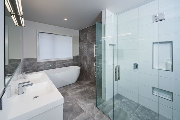 Bright contemporary bathroom with a freestanding white bathtub, double vanity, large glass-enclosed walk-in shower, white wall tiles, grey stone-look floor and wall tiles, and recessed ceiling lighting.