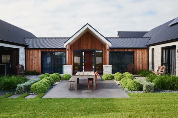 Single-storey courtyard-style residential building with a central gabled entry pavilion, dark metal pitched roofs, vertical timber cladding, and pale masonry side wings. Large glazed doors and windows open toward a paved courtyard furnished with an outdoor dining table and chairs, surrounded by clipped shrubs and ornamental grasses.