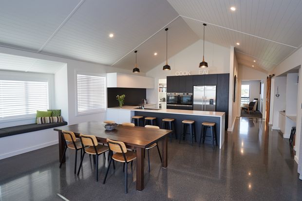 Open-plan kitchen and dining area with a dark timber dining table and chairs, a large island with bar stools, dark cabinetry, stainless-steel appliances, polished concrete flooring, white panelled ceilings, pendant lights, and a built-in window seat. A hallway and adjacent rooms are visible beyond the kitchen.