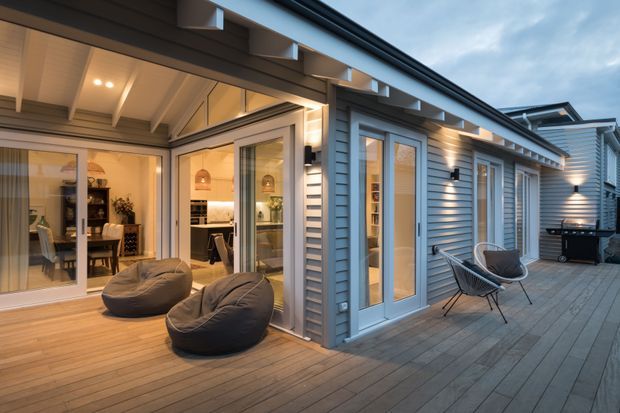 A contemporary covered timber deck and courtyard terrace wraps around a glazed extension, with large sliding and hinged glass doors opening into a warmly lit dining and kitchen area. Grey horizontal weatherboard cladding, white-framed glazing, exposed timber roof framing, integrated exterior lighting, outdoor lounge chairs, beanbags, and a barbecue define the space.