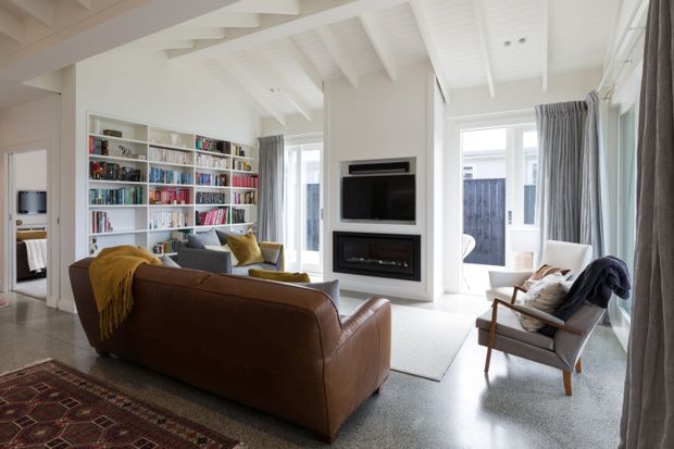 Bright, refined living room with a high white painted ceiling featuring exposed beams and tongue-and-groove detailing. The room includes a large built-in white bookcase filled with books, a wall-mounted television above a built-in fireplace, a brown leather sofa, upholstered chairs, layered rugs, and full-height grey curtains around generous windows and glazed doors.