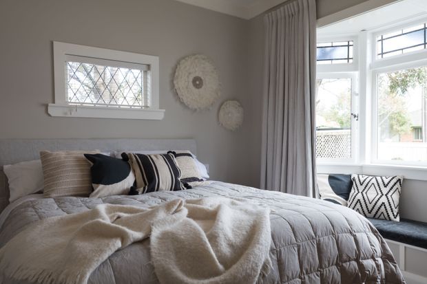A softly lit, inviting bedroom with a made bed layered in a grey quilt, cream throw, and patterned cushions. The room has warm grey walls, white trim, a small diamond-leaded window, full-height grey curtains, and a bright bay-style window with a built-in window seat. Neutral textiles and woven wall decor create a calm, cozy interior.