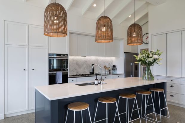 Bright, spacious contemporary kitchen with a large dark island and seating for four, white shaker-style cabinetry, integrated appliances, pale stone benchtops, a marble-look herringbone splashback, and three woven pendant lights beneath a white beamed ceiling.