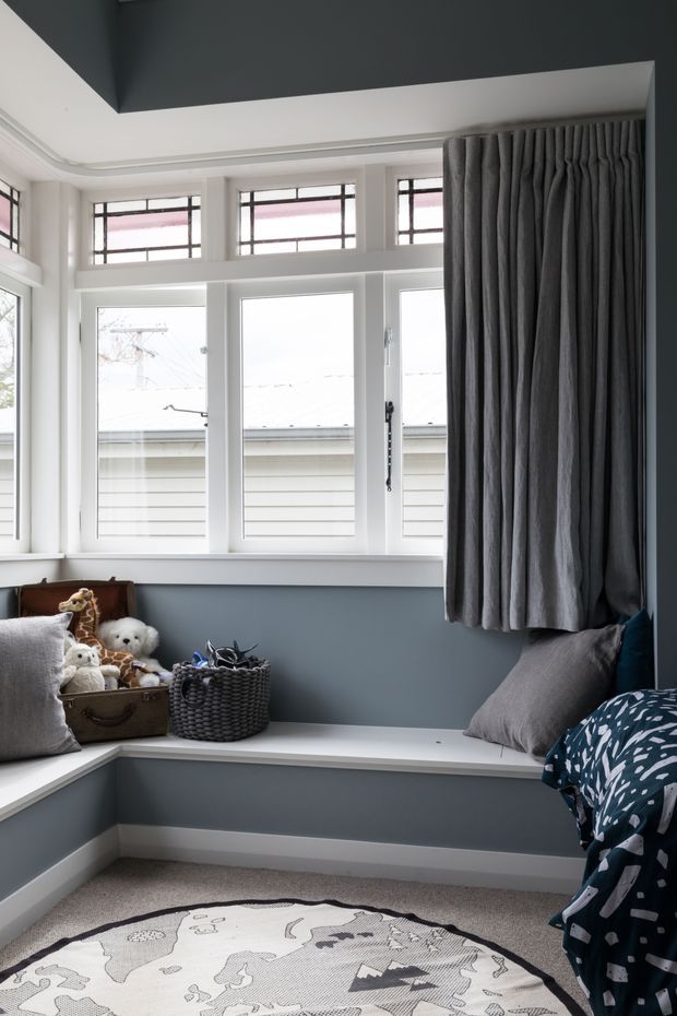 A cozy children's bedroom nook with a built-in white window seat wrapping around the corner beneath a bank of white-framed windows. The room has blue-grey walls, a dark charcoal ceiling recess, heavy charcoal curtains, soft grey cushions, plush toys, a woven basket, and a patterned duvet and round rug.