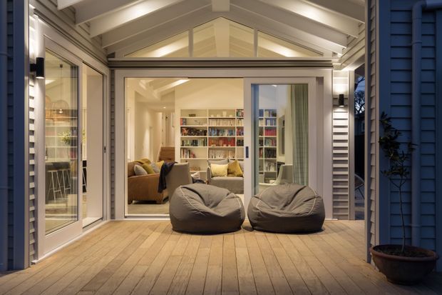 A covered timber deck or outdoor entertaining area opens directly into a warmly lit contemporary living room. Two large beanbag seats sit in the foreground, with glazed sliding doors, built-in bookshelves, a sofa, and a peaked glazed ceiling visible inside. The exterior facade uses dark blue weatherboards with white trim, while the deck and interior are finished in warm pale timber tones.