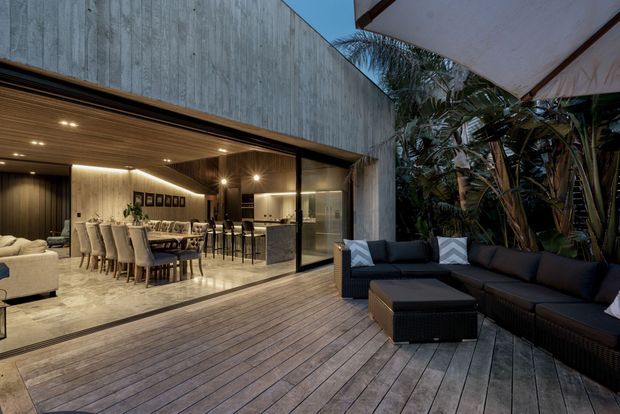 A sophisticated modern indoor-outdoor entertaining area opens from a large glazed living and dining space onto a spacious timber deck. The interior features a long dining table, upholstered dining chairs, a lounge area, a kitchen or bar counter, warm timber ceiling lining, and integrated lighting. Dark outdoor sectional seating and ottomans occupy the deck beside dense tropical planting, while a broad concrete-toned exterior wall frames the opening.