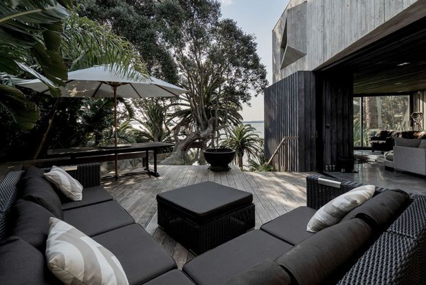 A sophisticated outdoor terrace adjoining a dark, timber-clad contemporary home. The spacious deck is furnished with black woven lounge seating, a low square ottoman, and a large neutral umbrella. Lush tropical and native planting surrounds the terrace, while an open glazed living area is visible to the right. The setting appears elevated, with a glimpse of water or distant landscape beyond the trees.