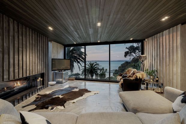 A sophisticated living room with full-height ocean-view glazing, vertical timber wall panelling, a dark timber-lined ceiling, and a linear fireplace. A large pale sectional sofa faces the fireplace and view, with a cowhide rug, sculptural side tables, and warm recessed lighting creating a luxurious coastal retreat atmosphere.