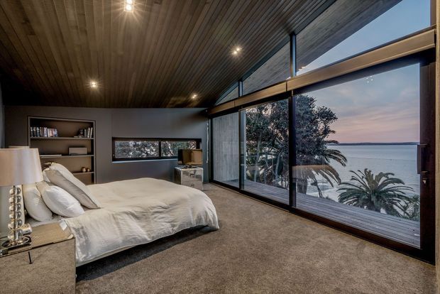 A spacious bedroom with a large bed, built-in shelving, dark wood-lined ceiling, grey walls, and carpeted flooring. Full-height glazing spans one side of the room, providing expansive coastal and ocean views with palm trees outside. The room has a warm, moody, contemporary feel with integrated ceiling lighting and dark window frames.
