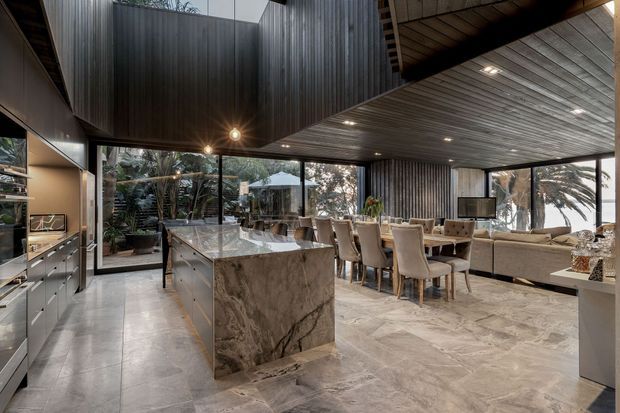 A luxurious open-plan kitchen, dining, and living area with floor-to-ceiling glazing opening toward a landscaped garden. The kitchen features a large veined stone island and dark built-in cabinetry, while the dining area has upholstered chairs beneath a timber-lined ceiling. Dark ribbed wall surfaces and exposed timber ceiling elements create a dramatic contemporary interior, complemented by pale stone flooring and extensive glazing.
