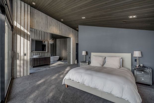A sophisticated contemporary bedroom with a large upholstered bed, white bedding, matching bedside tables and table lamps. The room features dark grey carpet, a charcoal-grey wall behind the bed, and dramatic vertically ribbed timber or wood-look wall and ceiling panels. A television and freestanding bathtub are visible through an open partition, suggesting an integrated bedroom and bathroom arrangement. Strong natural light enters from full-height glazing on the left.