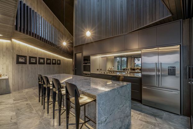A spacious, dark-toned contemporary kitchen with a large stone island and seating for four. Full-height timber wall and ceiling panelling create a dramatic interior, complemented by integrated dark cabinetry, a stainless-steel refrigerator, under-cabinet lighting, pendant bulbs, and a tiled floor. A high-level void or mezzanine-like opening with vertical timber screening adds architectural interest.