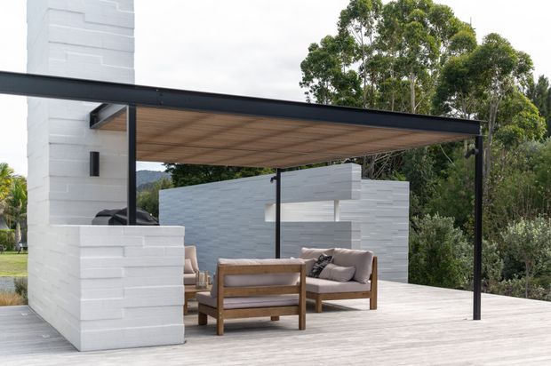 Contemporary outdoor entertaining area with a flat black-framed canopy, timber-lined ceiling, pale timber decking, white painted brick walls and a built-in fireplace or chimney structure. A timber outdoor lounge setting sits beneath the covered terrace, surrounded by established garden vegetation.