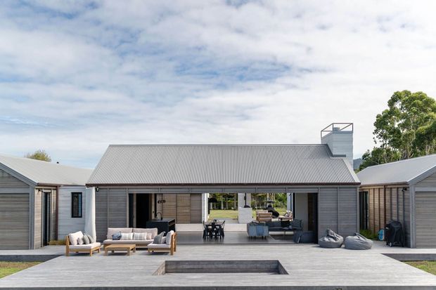 A wide exterior view of a contemporary single-storey residence arranged around a broad central outdoor entertaining area. The home has grey horizontal cladding, corrugated pitched roofs, extensive central glazing and doors, a covered alfresco zone with outdoor seating and dining, and a sunken rectangular pool or water feature in the foreground. A rooftop chimney or vent structure is visible above the main roof.